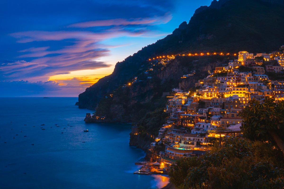 Positano oplyst i tusmørket — farverige bygninger lysende mod klippen over havet.